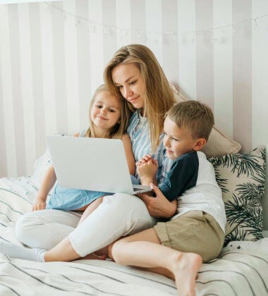 A woman and her two children use a laptop whilst sitting on a bed